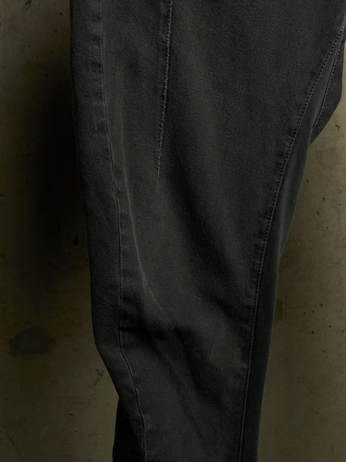 A close-up of dark charcoal gray denim pants, showing the fabric texture, seam details, and a slight fade on the thigh, set against a dark concrete background.