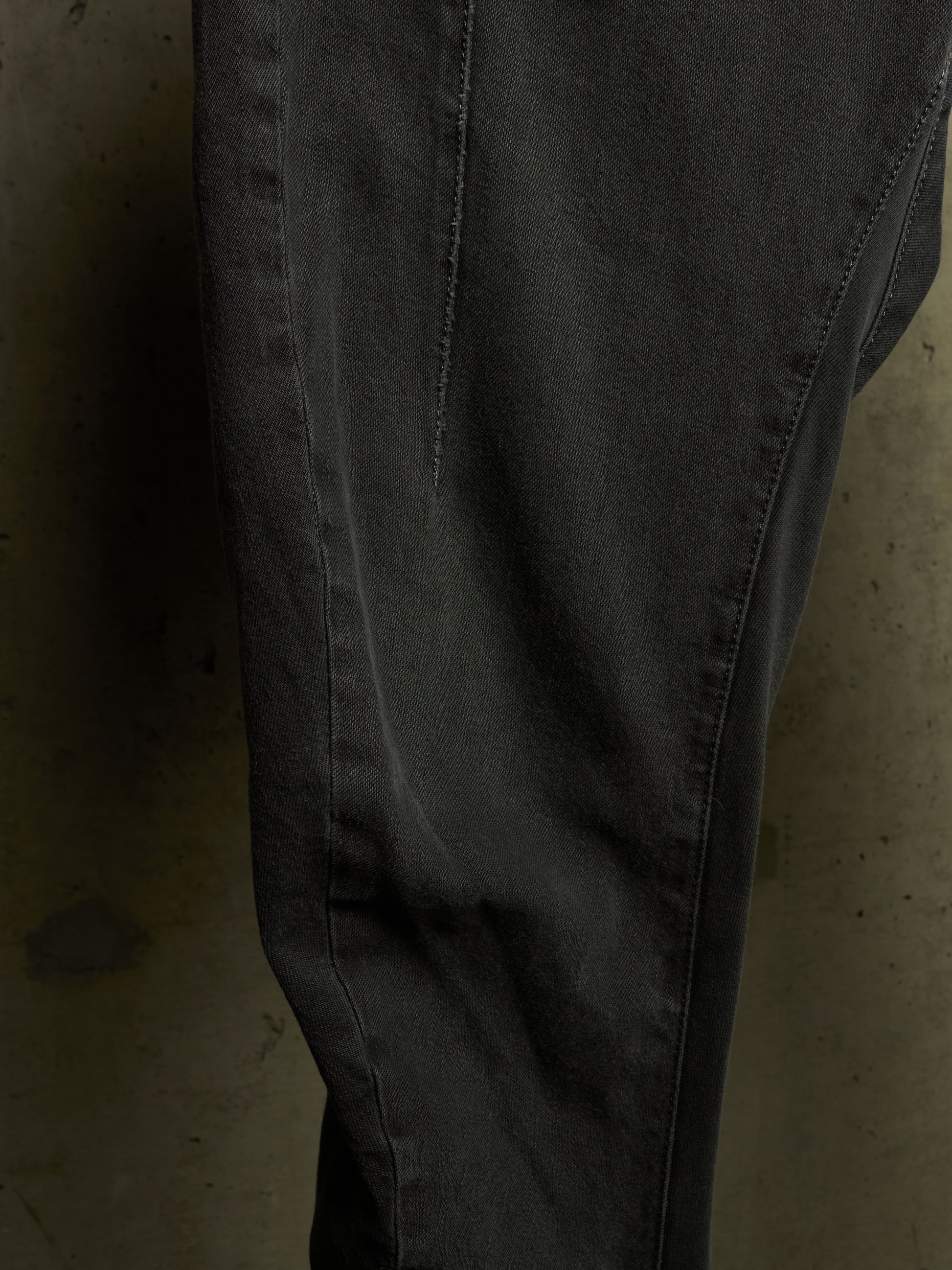 A close-up of dark charcoal gray denim pants, showing the fabric texture, seam details, and a slight fade on the thigh, set against a dark concrete background.