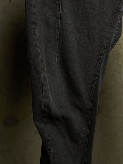A close-up of dark charcoal gray denim pants, showing the fabric texture, seam details, and a slight fade on the thigh, set against a dark concrete background.