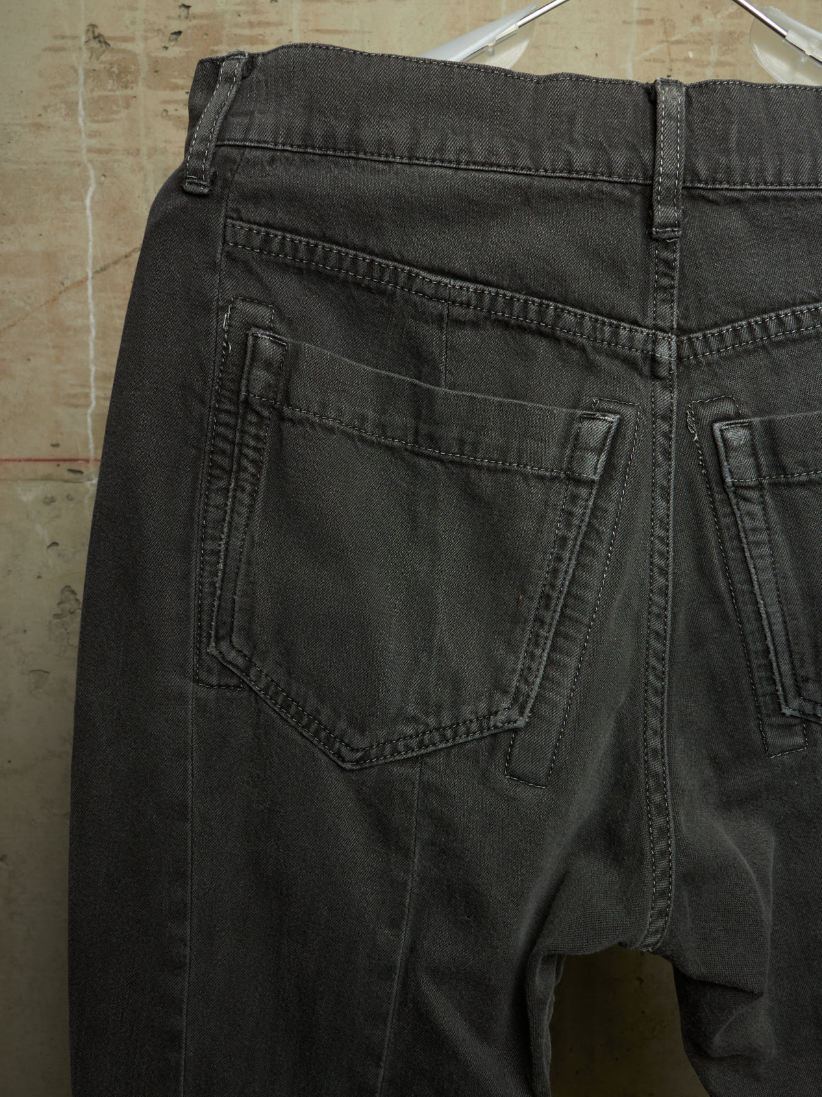 Close-up of the back waistband and rear pockets of dark charcoal gray denim jeans hanging against a textured beige wall.