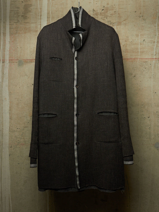 A dark charcoal gray wool-blend coat with contrasting light gray and white striped trim, hanging against a textured concrete wall.