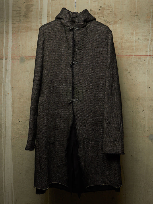 A dark brown textured wool coat with a high collar and toggle closures, hanging against a textured concrete wall.