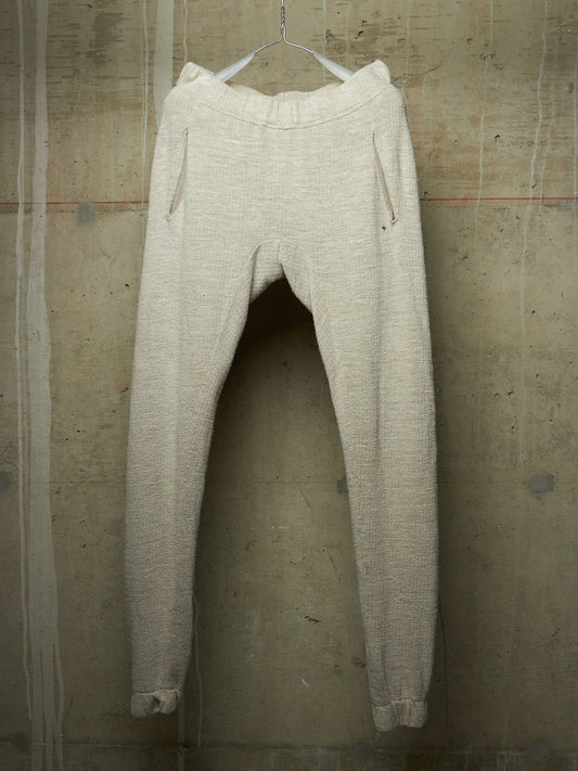A pair of light beige textured knit pants hanging from a metal hanger against a distressed concrete wall.