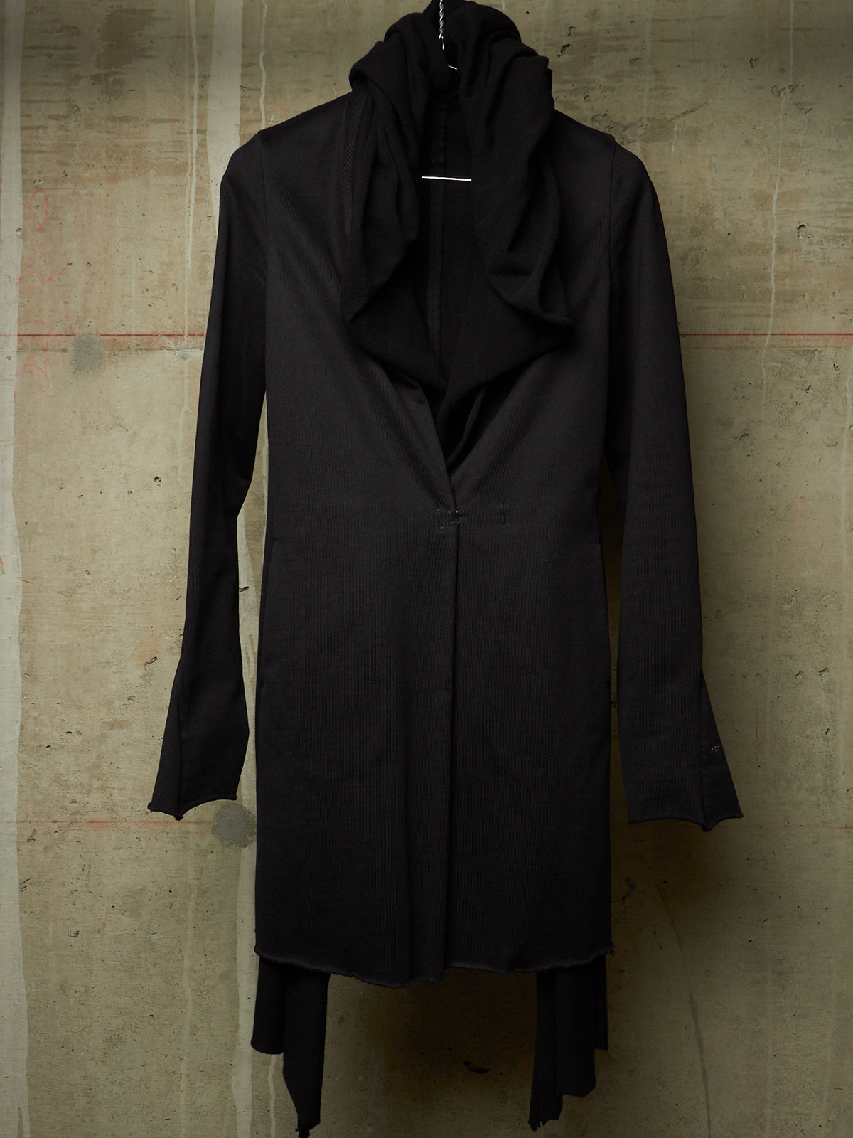 A long black draped coat with a high draped collar and long sleeves, displayed on a hanger against a textured concrete wall.