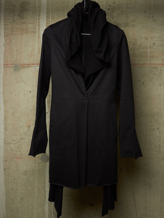 A long black draped coat with a high draped collar and long sleeves, displayed on a hanger against a textured concrete wall.