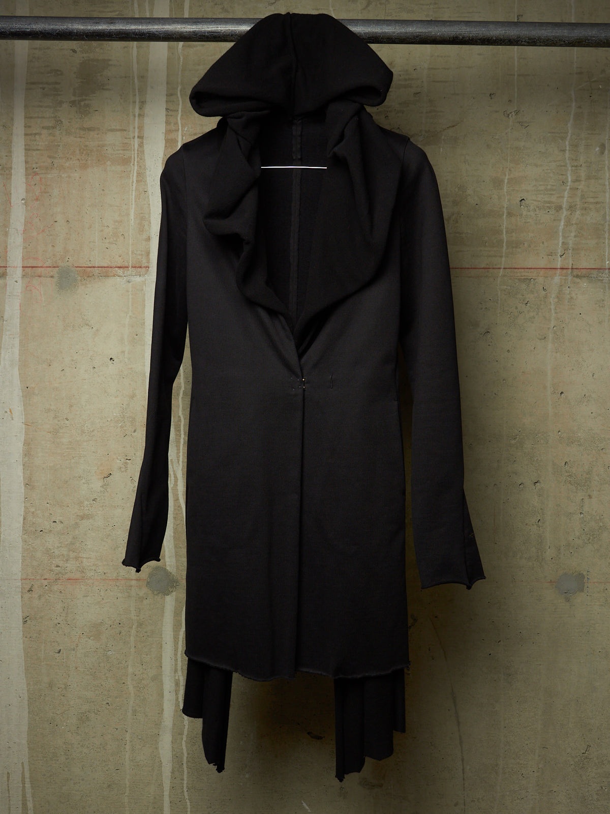 A long black hooded coat with a draped, asymmetrical front and long sleeves, hanging against a textured concrete wall.