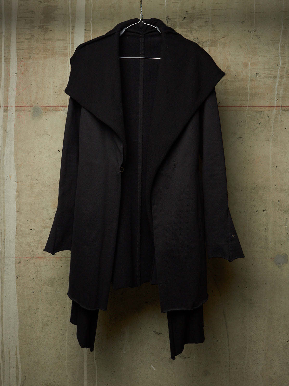 A black asymmetrical long coat with a wide draped collar, hanging on a wire hanger against a textured concrete wall.