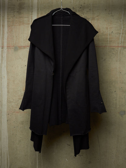 A black asymmetrical long coat with a wide draped collar, hanging on a wire hanger against a textured concrete wall.