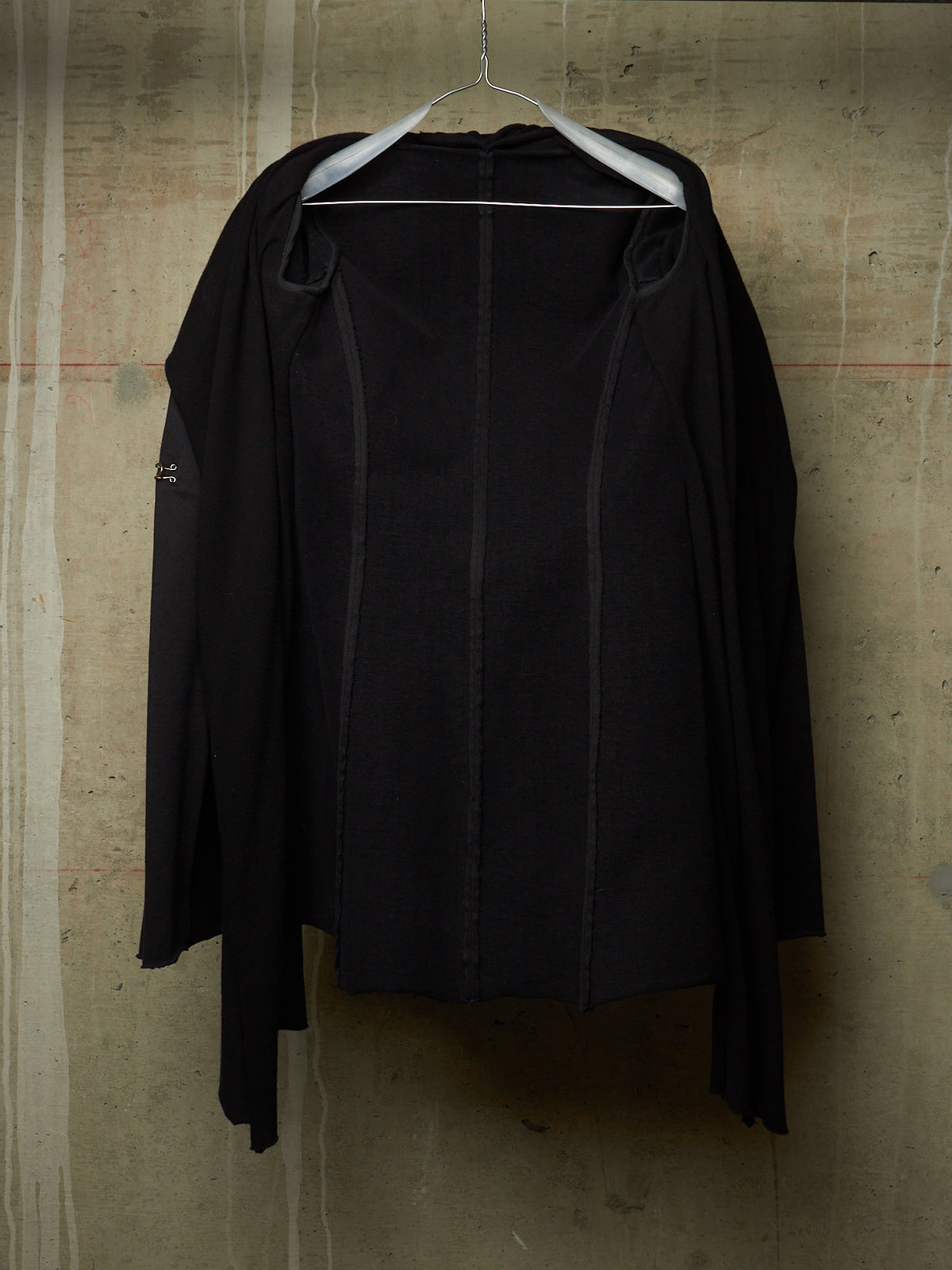 A black long-sleeve draped jacket or cardigan hanging on a white hanger against a textured concrete wall.