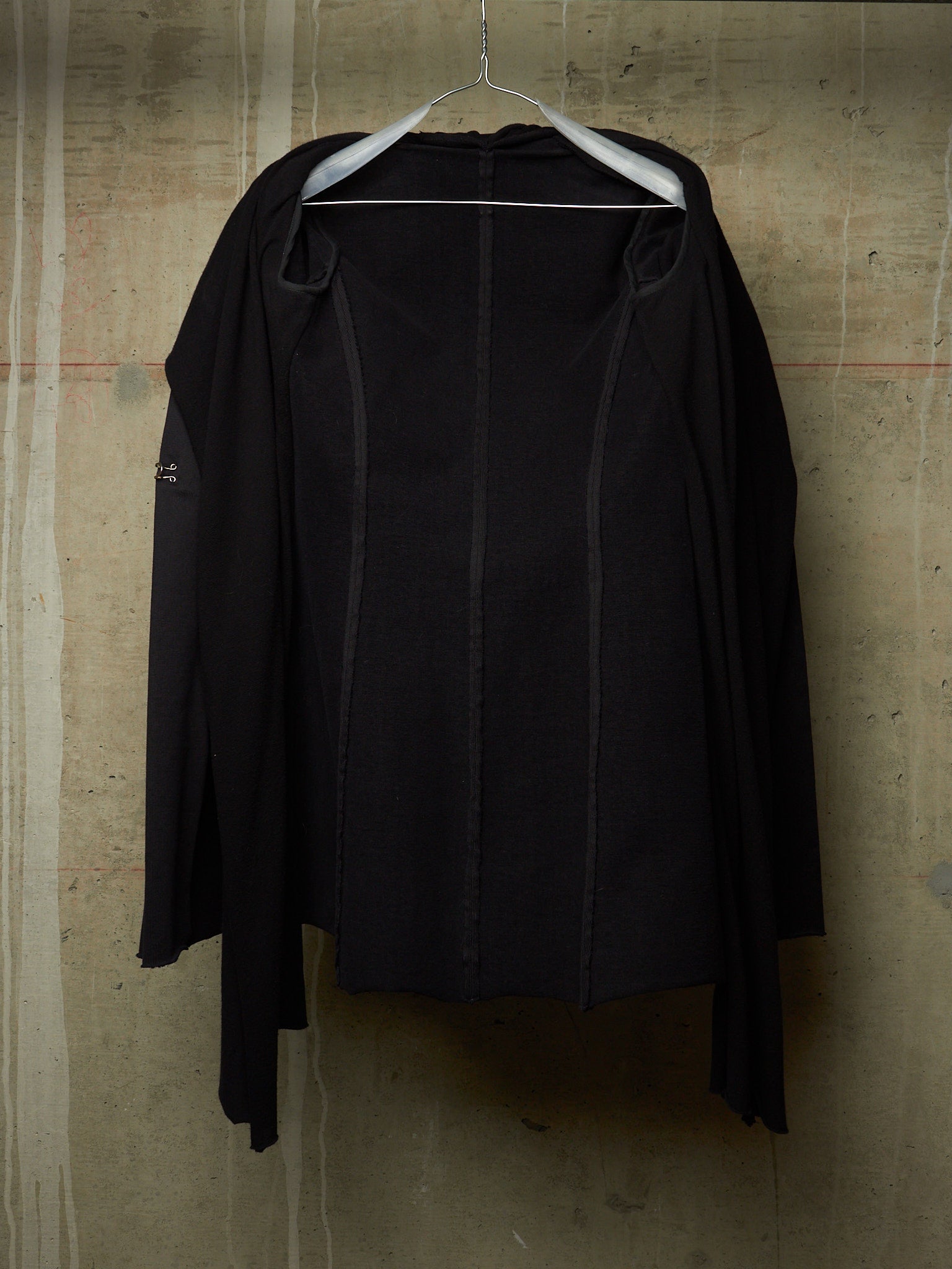 A black long-sleeve draped jacket or cardigan hanging on a white hanger against a textured concrete wall.