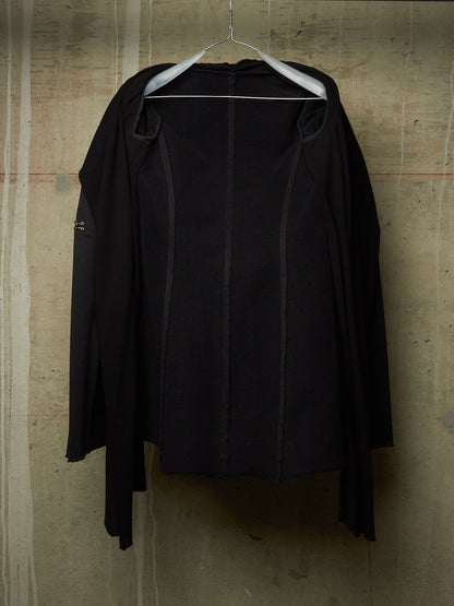 A black long-sleeve draped jacket or cardigan hanging on a white hanger against a textured concrete wall.