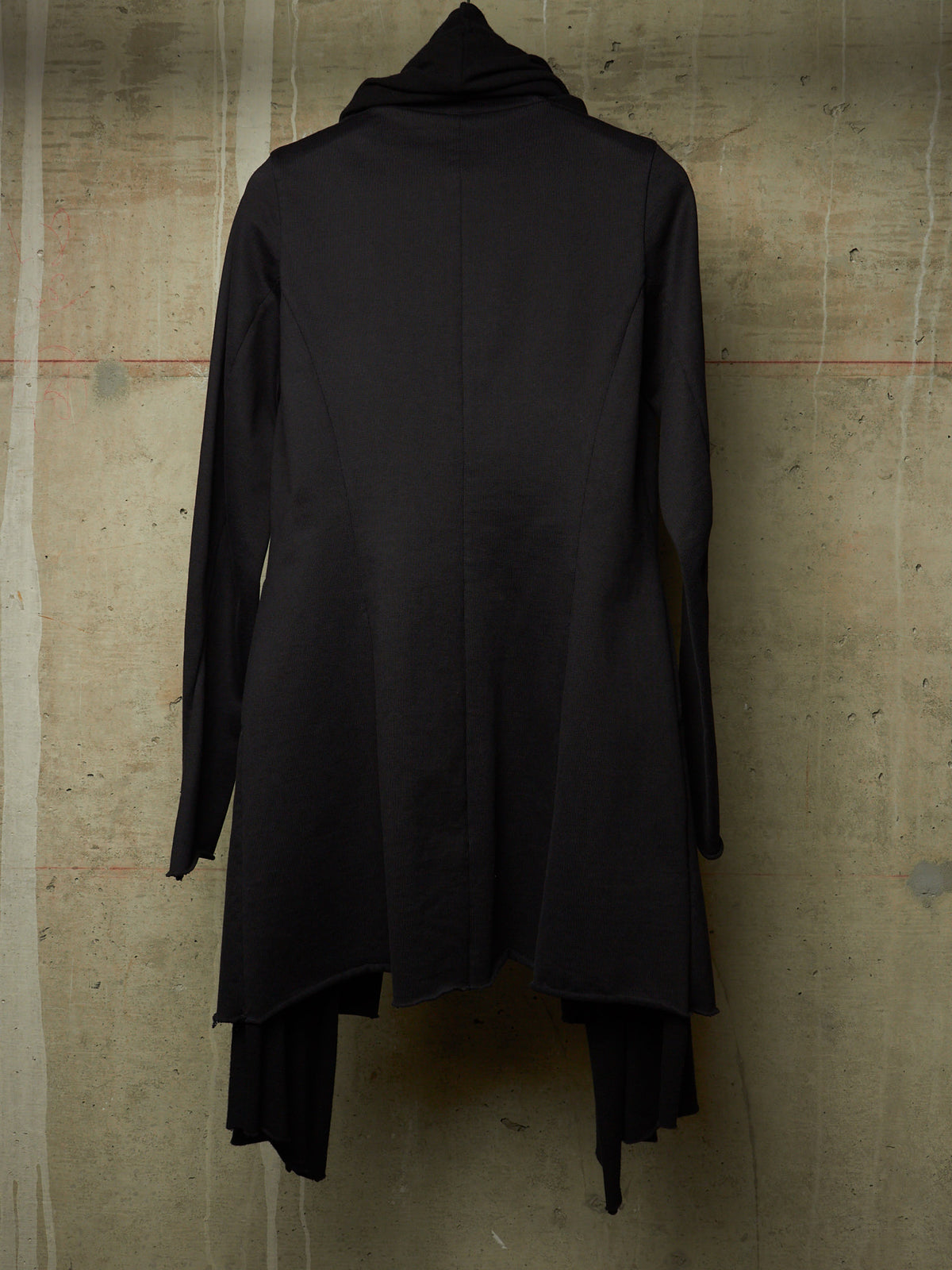 A black long-sleeve tunic top with a high draped neckline and an asymmetrical layered hem, displayed against a textured concrete wall.