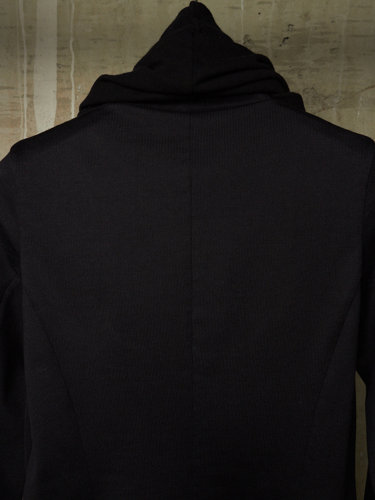 A close-up back view of a black hooded garment, likely a hoodie or long-sleeve top, shown against a textured beige wall.