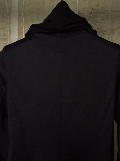 A close-up back view of a black hooded garment, likely a hoodie or long-sleeve top, shown against a textured beige wall.