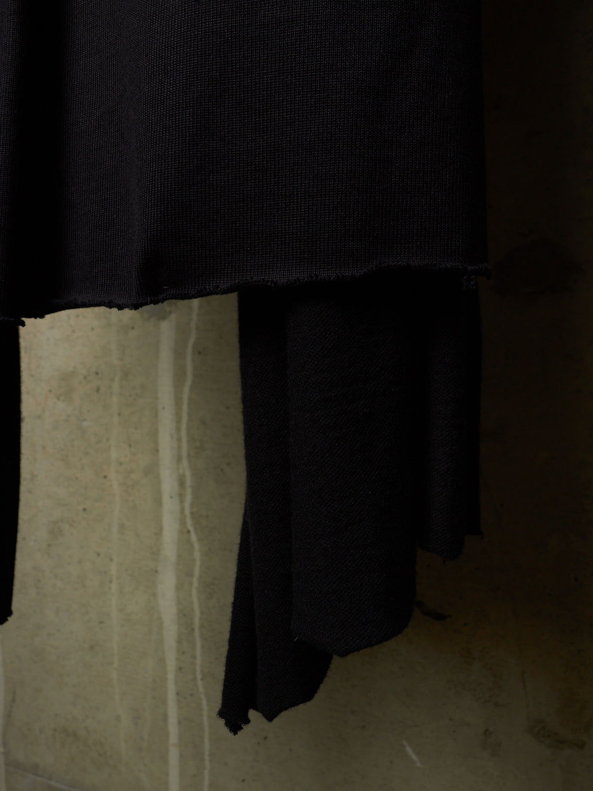 A close-up of a black ribbed knit fabric, likely a sweater or garment, draped over a textured concrete surface in a dimly lit setting.
