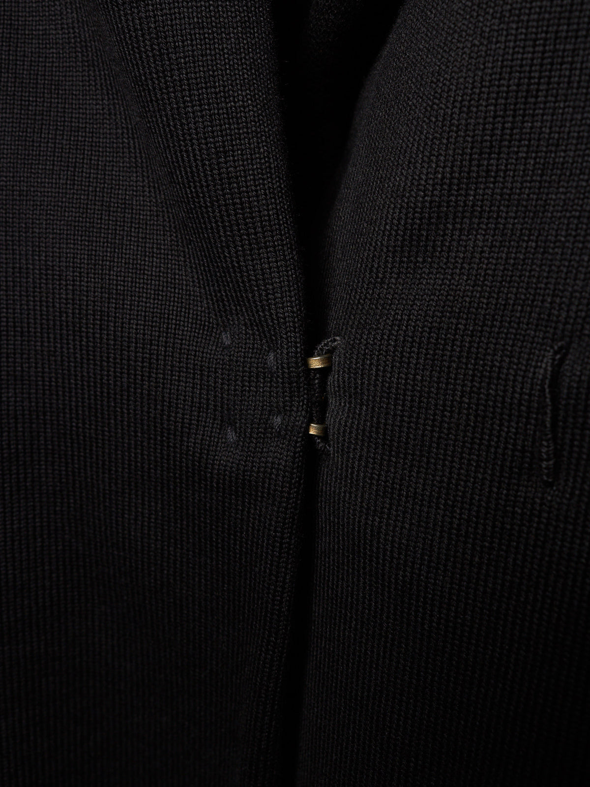 Close-up of a black textured knit garment, showing the fine ribbed fabric and a small gold-toned zipper pull.