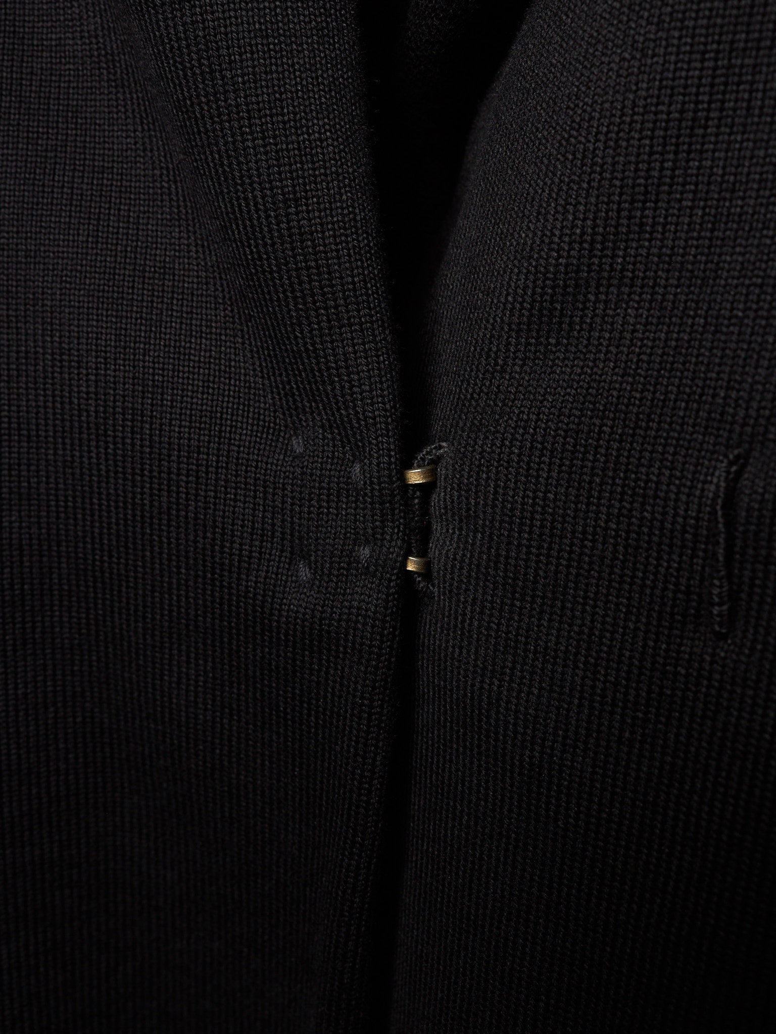 Close-up of a black textured knit garment, showing the fine ribbed fabric and a small gold-toned zipper pull.