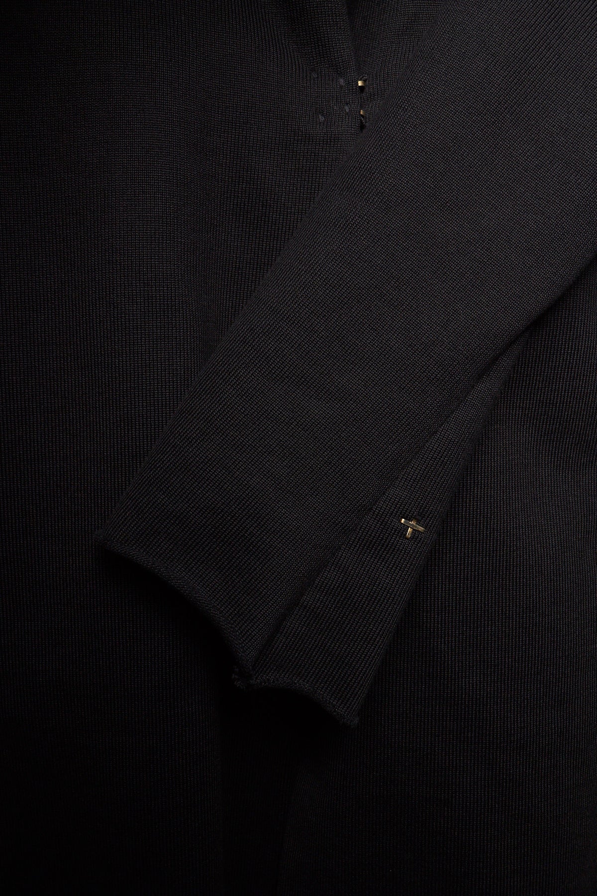 Close-up of a black ribbed knit garment, showing the texture of the fabric and a small gold embroidered logo on the cuff.