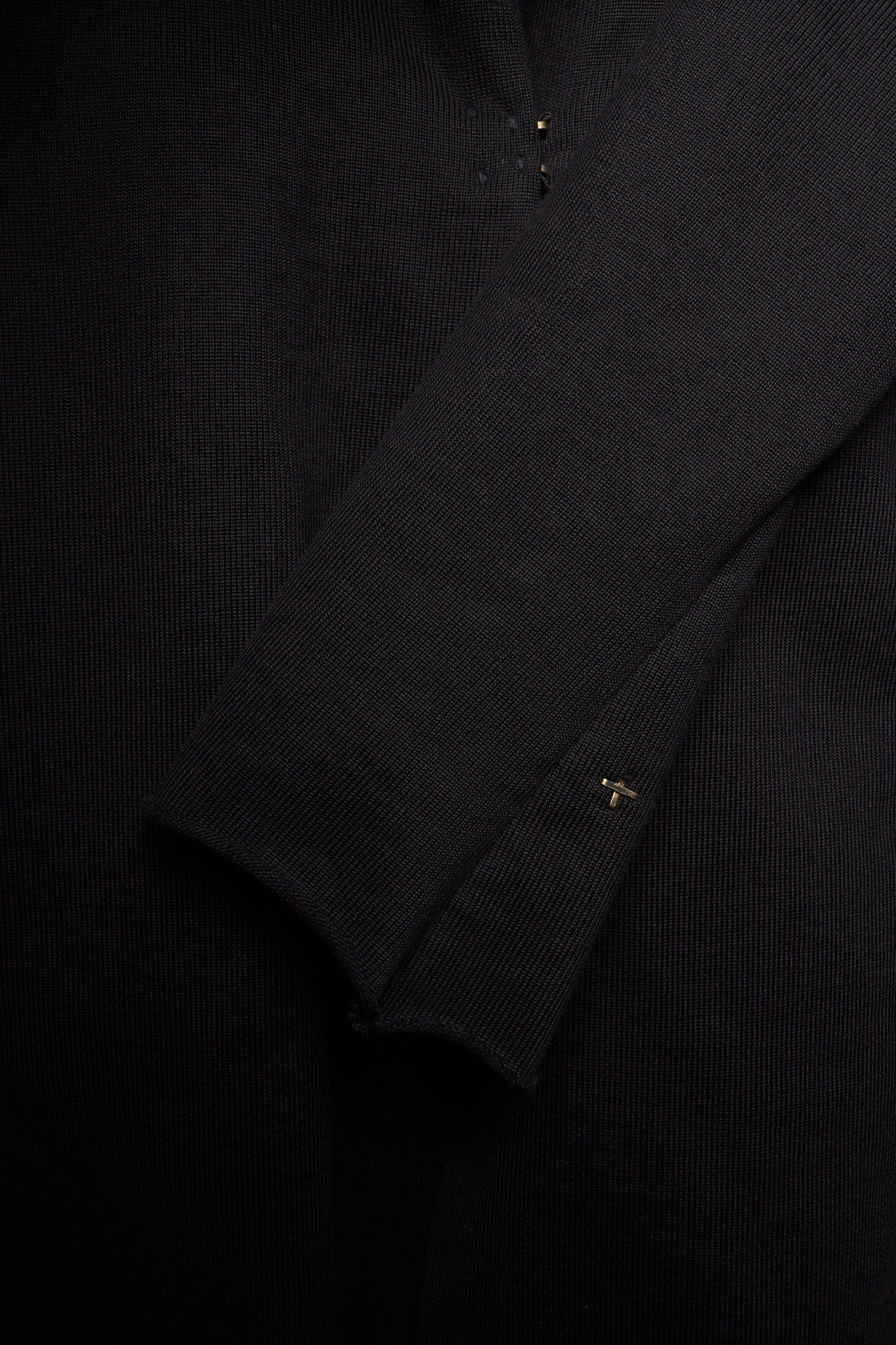 Close-up of a black ribbed knit garment, showing the texture of the fabric and a small gold embroidered logo on the cuff.