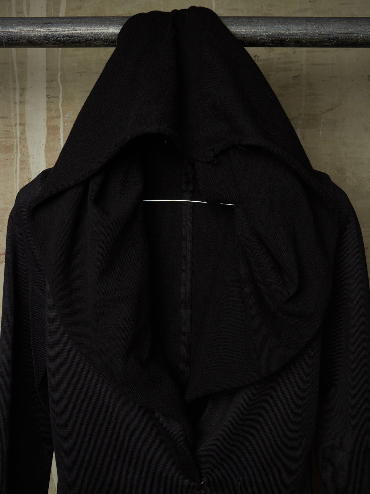 A black draped hooded garment or shawl hanging from a metal rod against a textured beige wall.