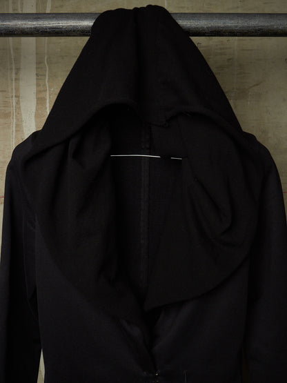 A black draped hooded garment or shawl hanging from a metal rod against a textured beige wall.