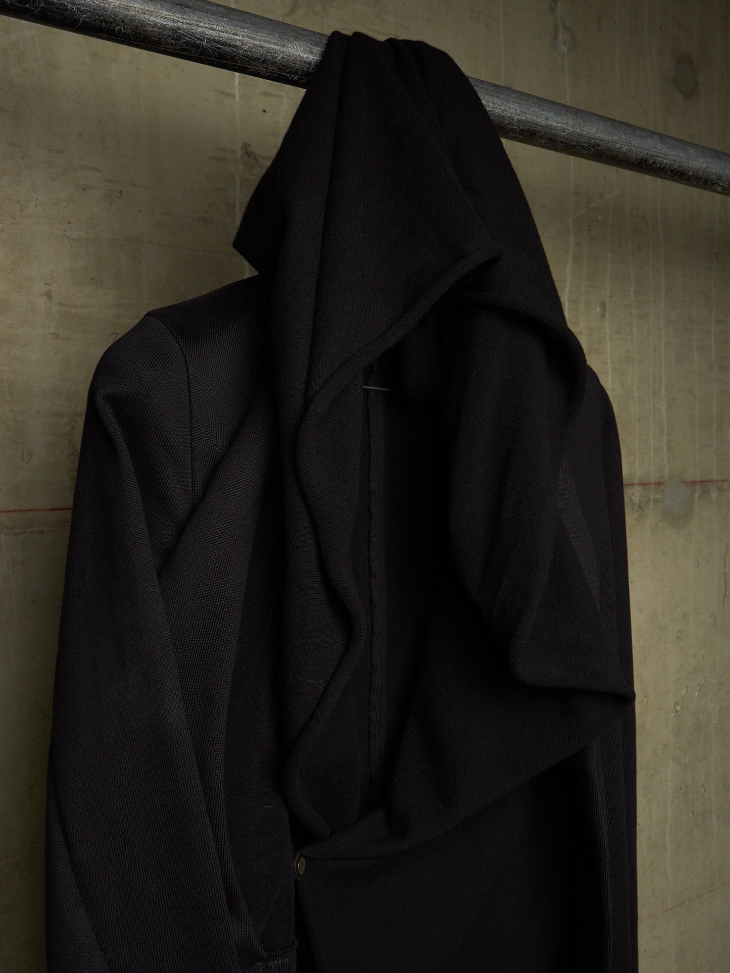 A black draped coat or wrap hanging from a metal rod against a textured concrete wall.