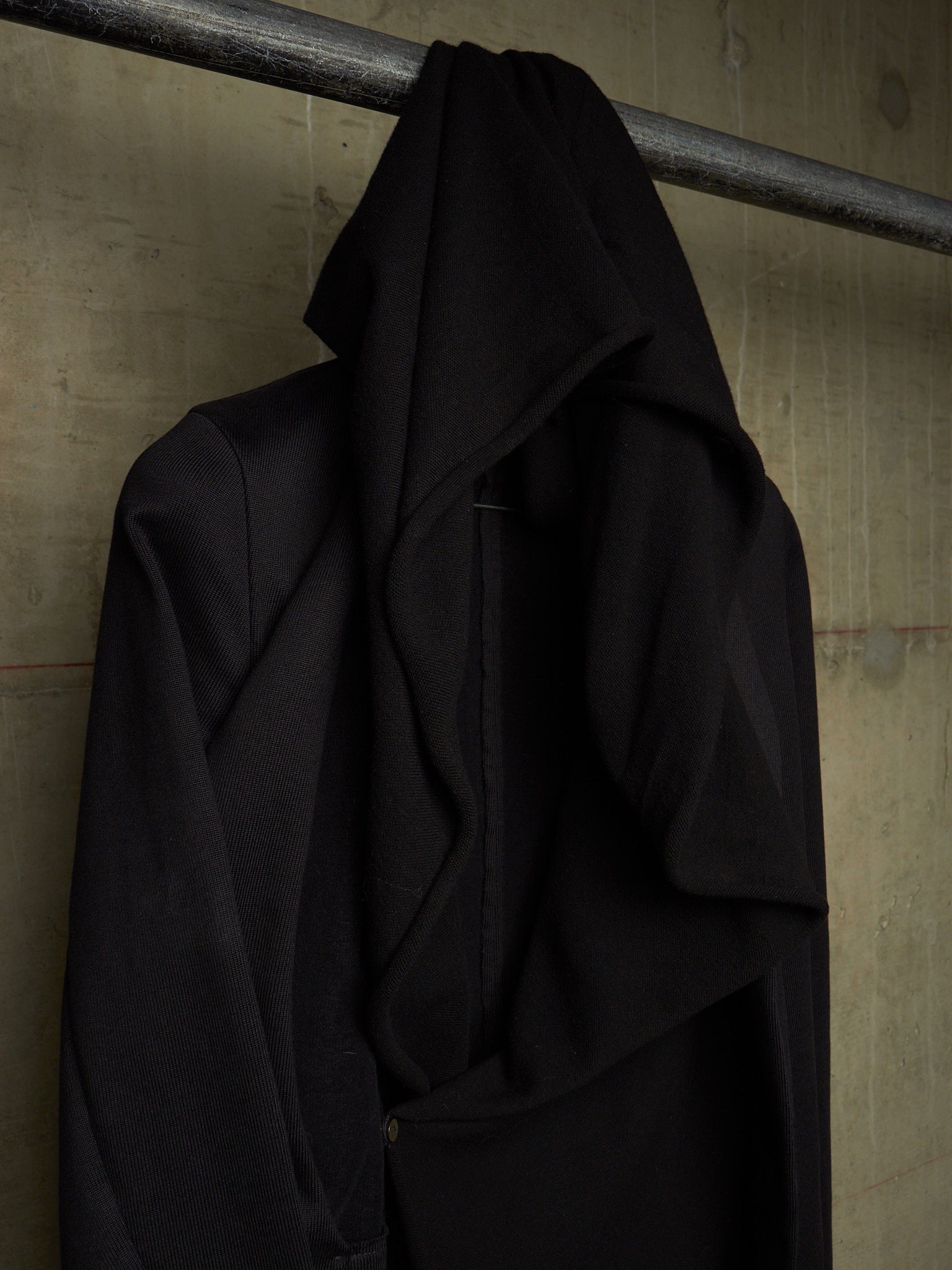 A black draped coat or wrap hanging from a metal rod against a textured concrete wall.