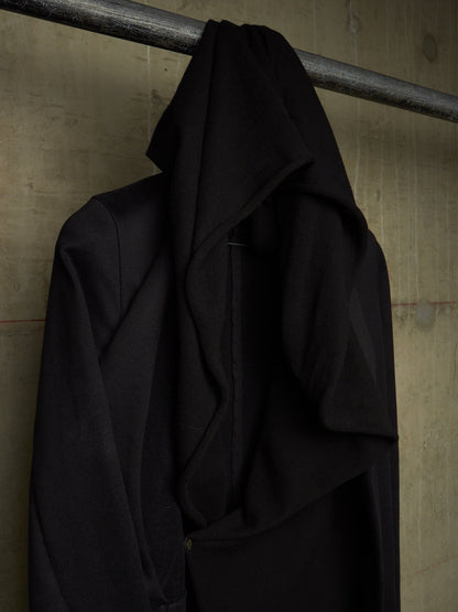 A black draped coat or wrap hanging from a metal rod against a textured concrete wall.