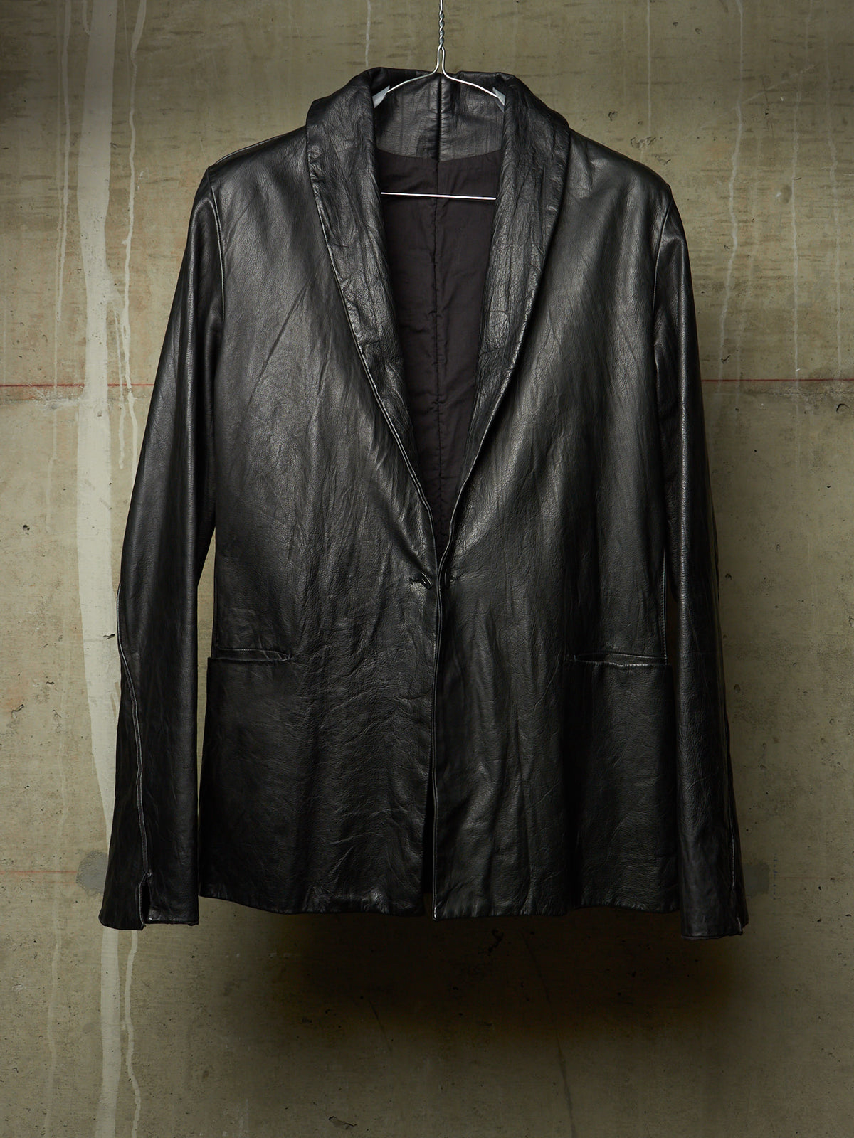 A black leather jacket with a shawl collar, hanging on a wire hanger against a textured concrete wall.