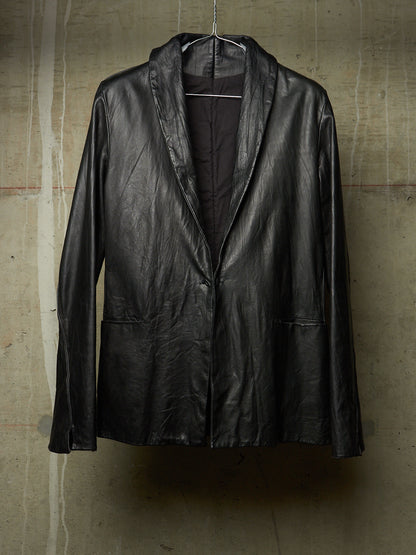 A black leather jacket with a shawl collar, hanging on a wire hanger against a textured concrete wall.