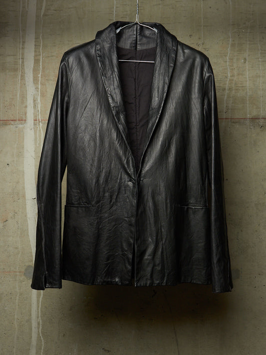 A black leather jacket with a shawl collar, hanging on a wire hanger against a textured concrete wall.