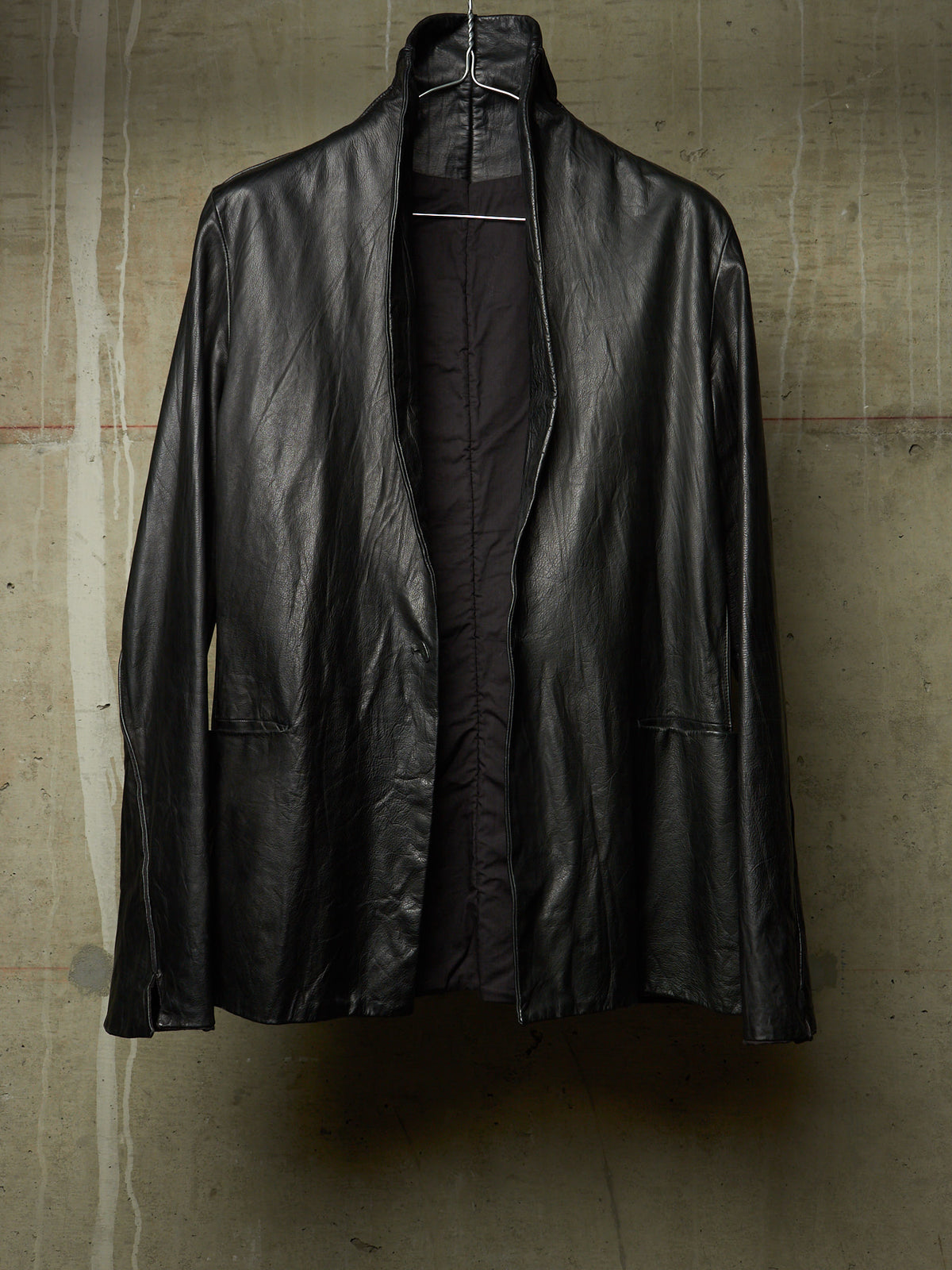 A black leather jacket with a high stand collar, hanging on a wire hanger against a textured concrete wall.