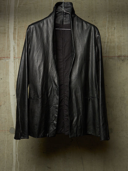 A black leather jacket with a high stand collar, hanging on a wire hanger against a textured concrete wall.