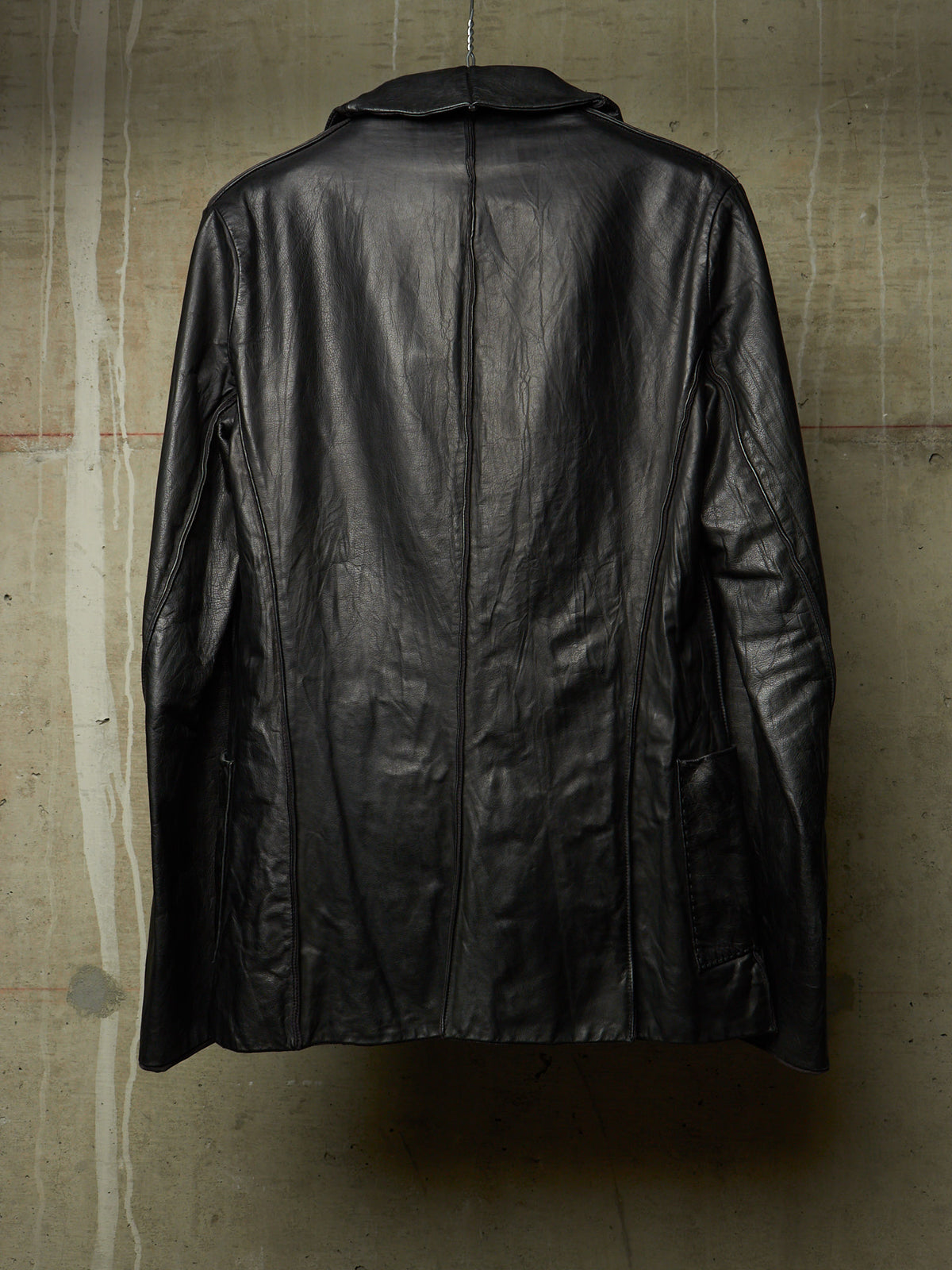 A black leather jacket hanging against a textured concrete wall, showing the back view with a central seam and collar.