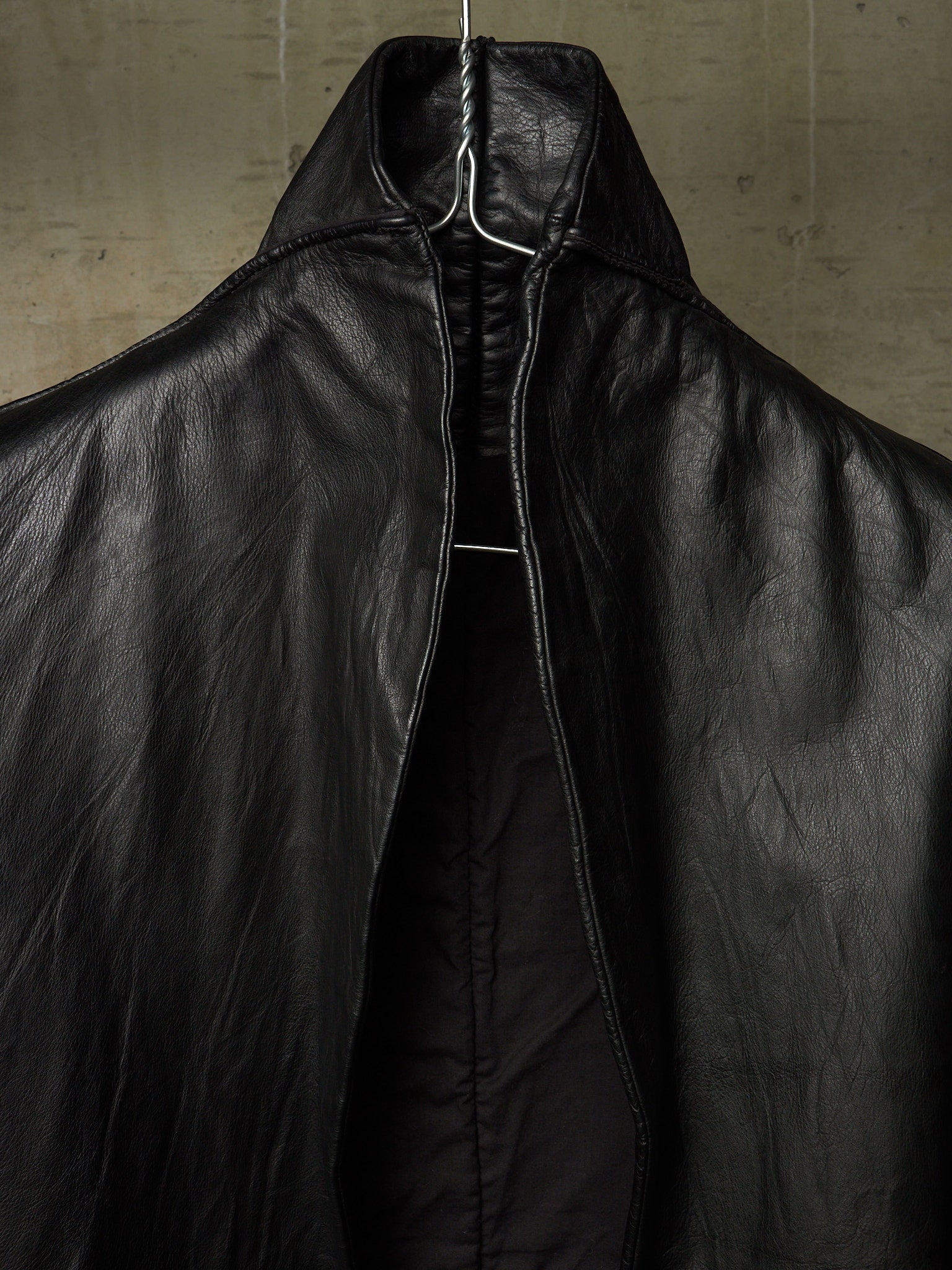 A close-up of a black leather jacket with a high collar and front zipper, hanging on a wire hanger against a textured concrete wall.
