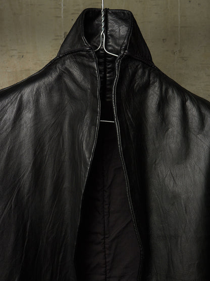 A close-up of a black leather jacket with a high collar and front zipper, hanging on a wire hanger against a textured concrete wall.