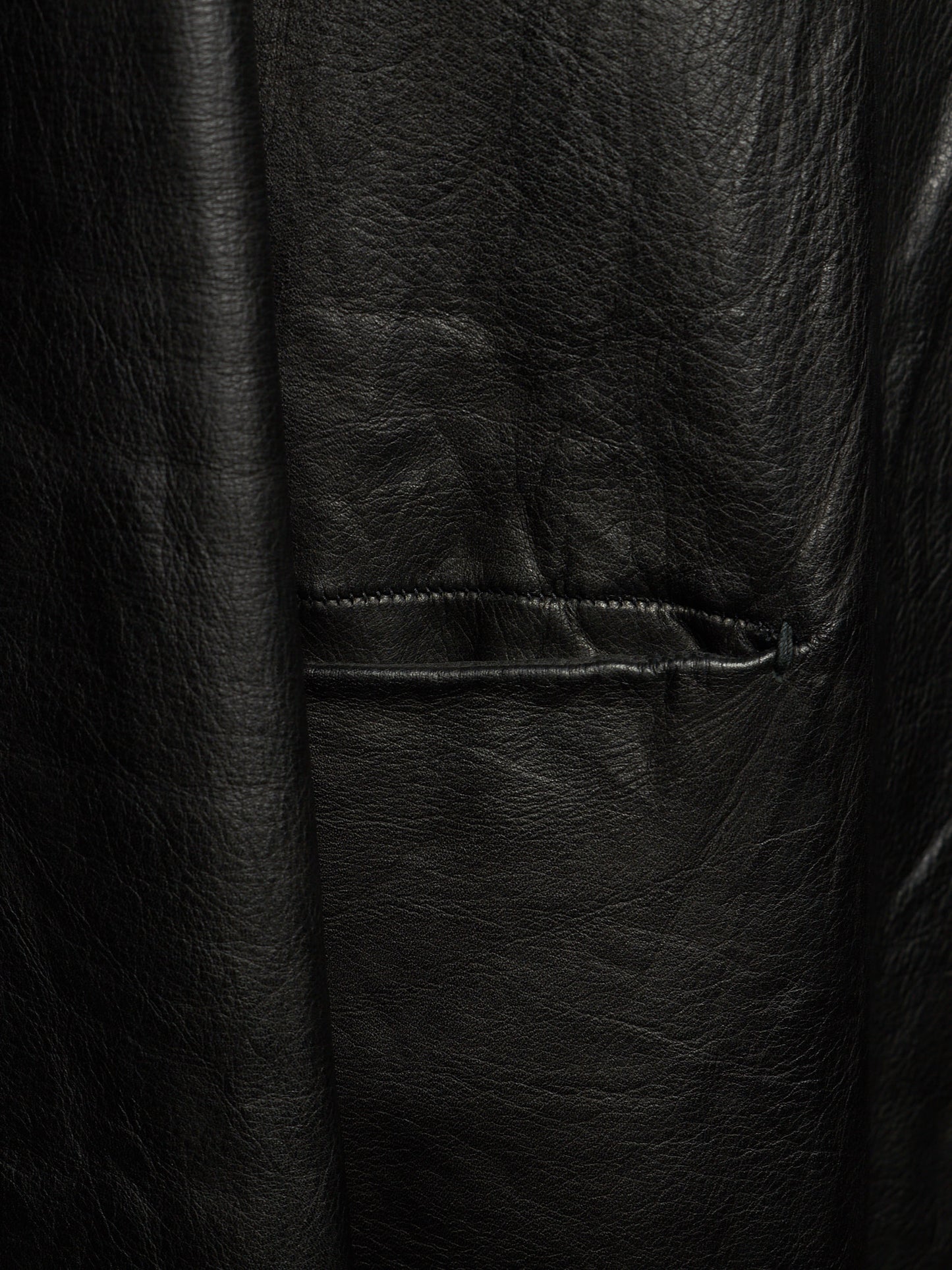 Close-up of a black leather jacket showing the textured grain and a horizontal welt pocket seam.