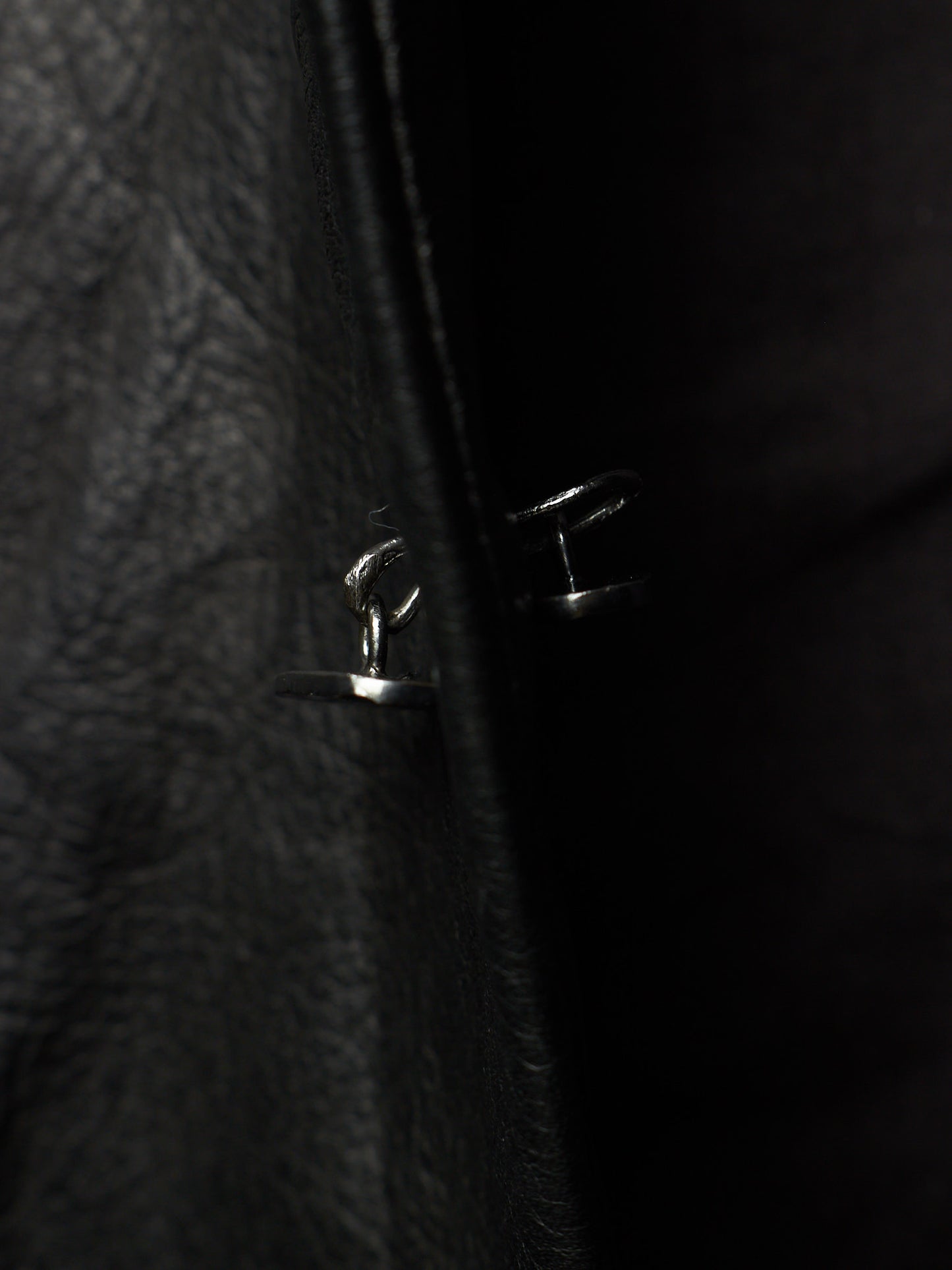 Close-up of a black leather jacket with a silver metal snap button closure, photographed in very dark lighting.