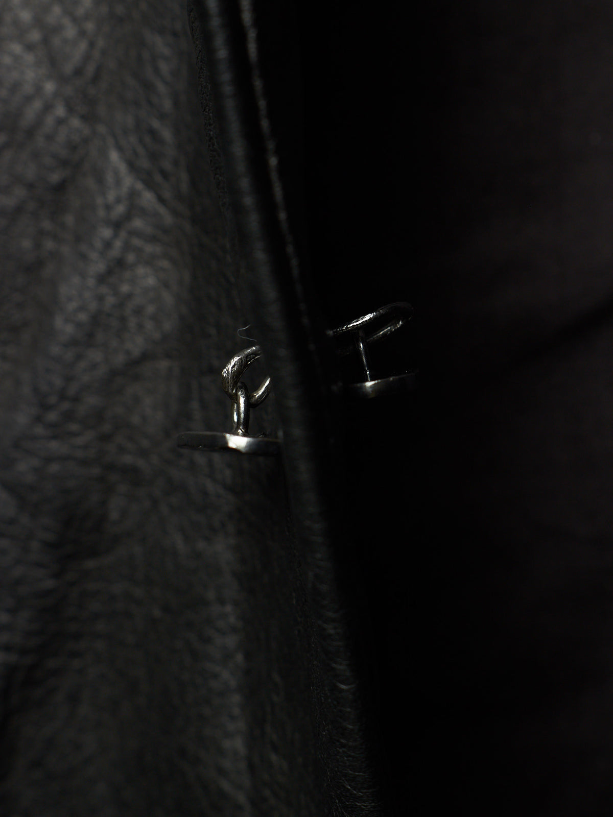 Close-up of a black leather jacket with a silver metal snap button closure, photographed in very dark lighting.