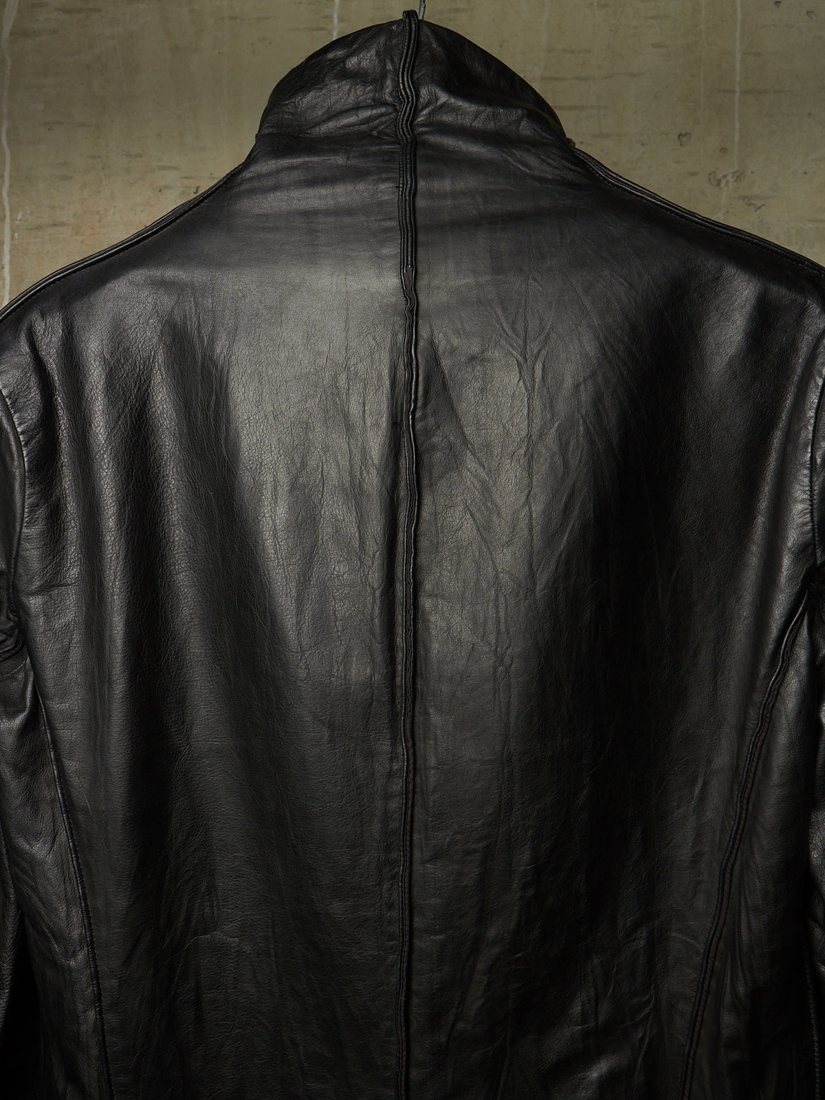 Close-up of the back of a black leather jacket with a high collar and a central seam, set against a textured beige background.