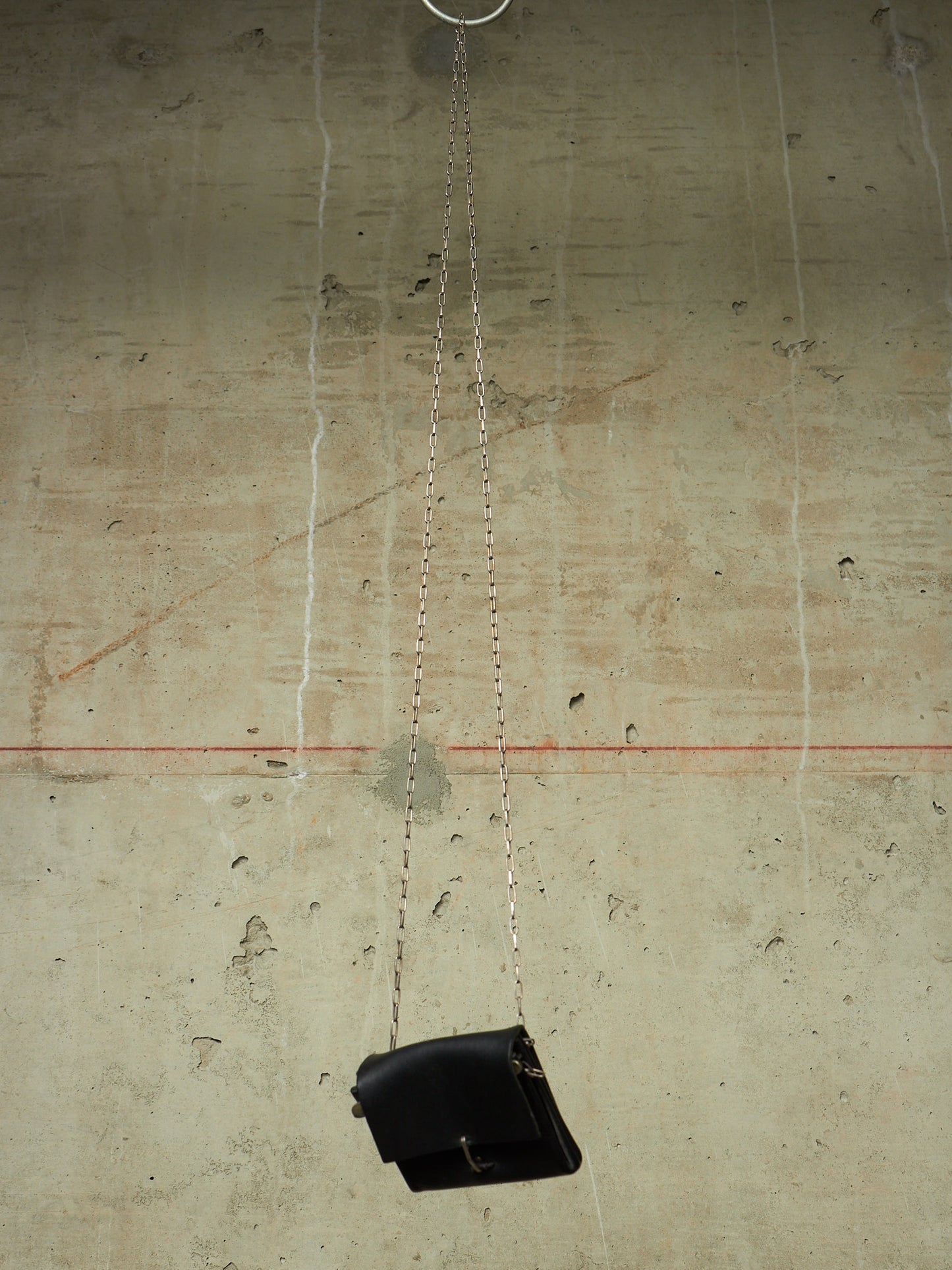 A black leather shoulder bag with a silver chain strap hangs from a metal ring against a textured concrete wall.