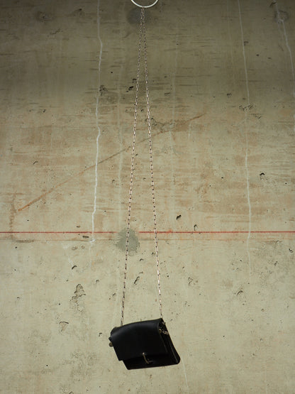 A black leather shoulder bag with a silver chain strap hangs from a metal ring against a textured concrete wall.