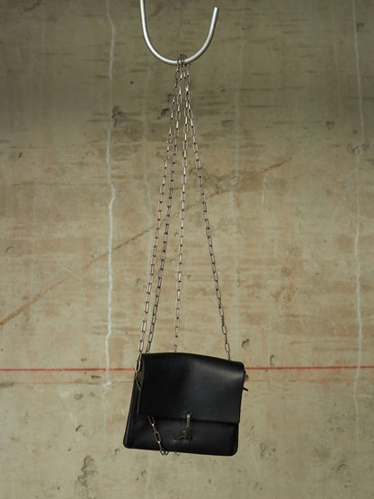 A black leather shoulder bag with a silver chain strap hangs from a metal hook against a textured concrete wall.