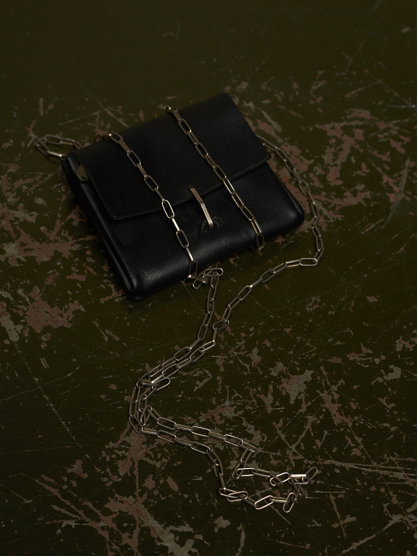 A black leather clutch bag with a gold-tone chain strap and a small gold clasp, photographed on a dark textured surface.