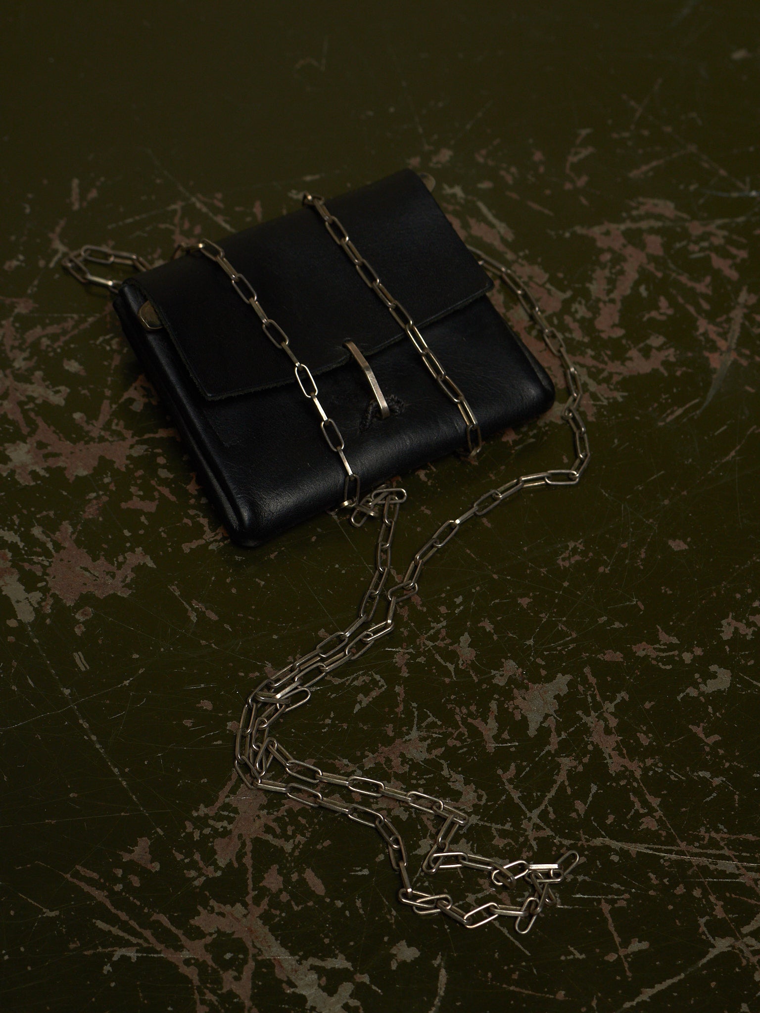 A black leather clutch bag with a gold-tone chain strap and a small gold clasp, photographed on a dark textured surface.