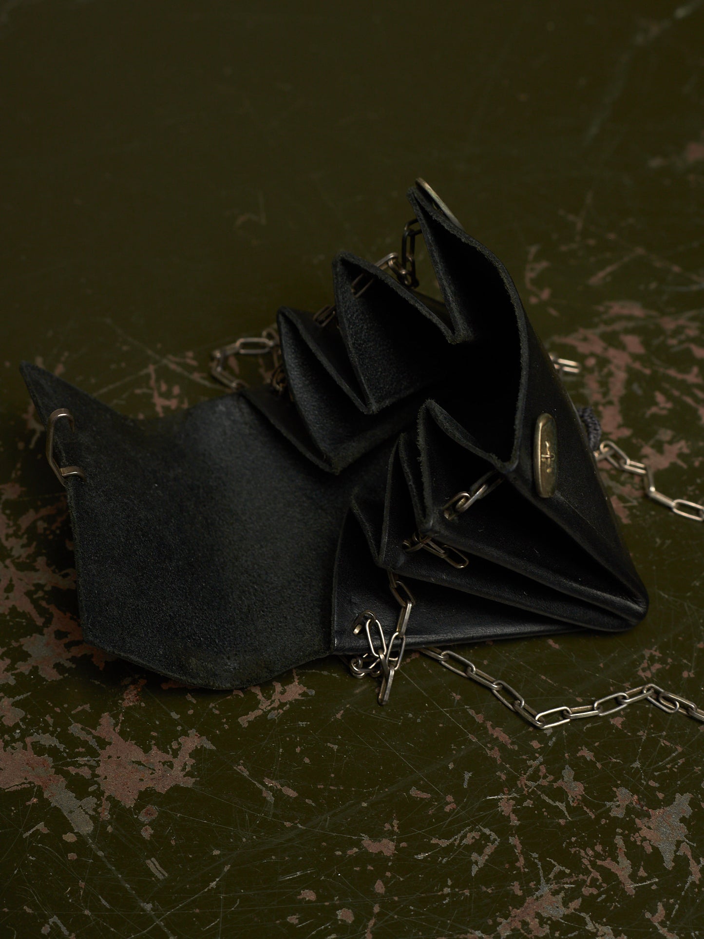 A black suede-like accordion-style clutch or wallet with silver-tone chain straps and a small oval metal clasp, photographed on a dark distressed surface.