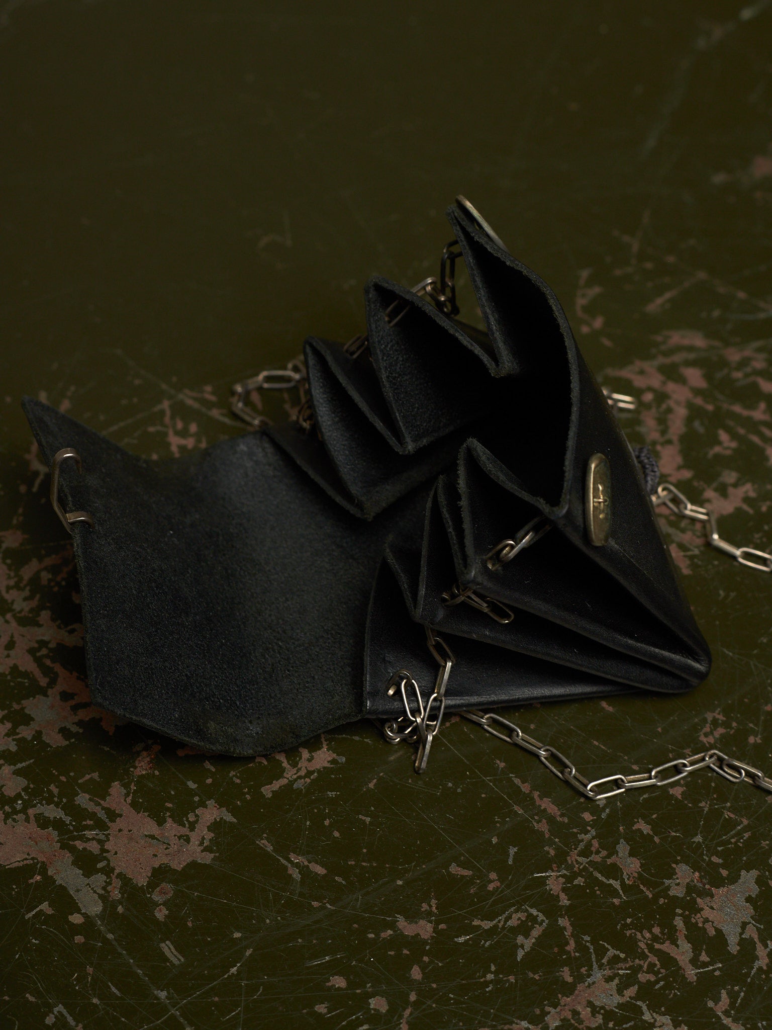 A black suede-like accordion-style clutch or wallet with silver-tone chain straps and a small oval metal clasp, photographed on a dark distressed surface.