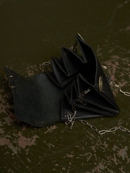 A black suede-like accordion-style clutch or wallet with silver-tone chain straps and a small oval metal clasp, photographed on a dark distressed surface.