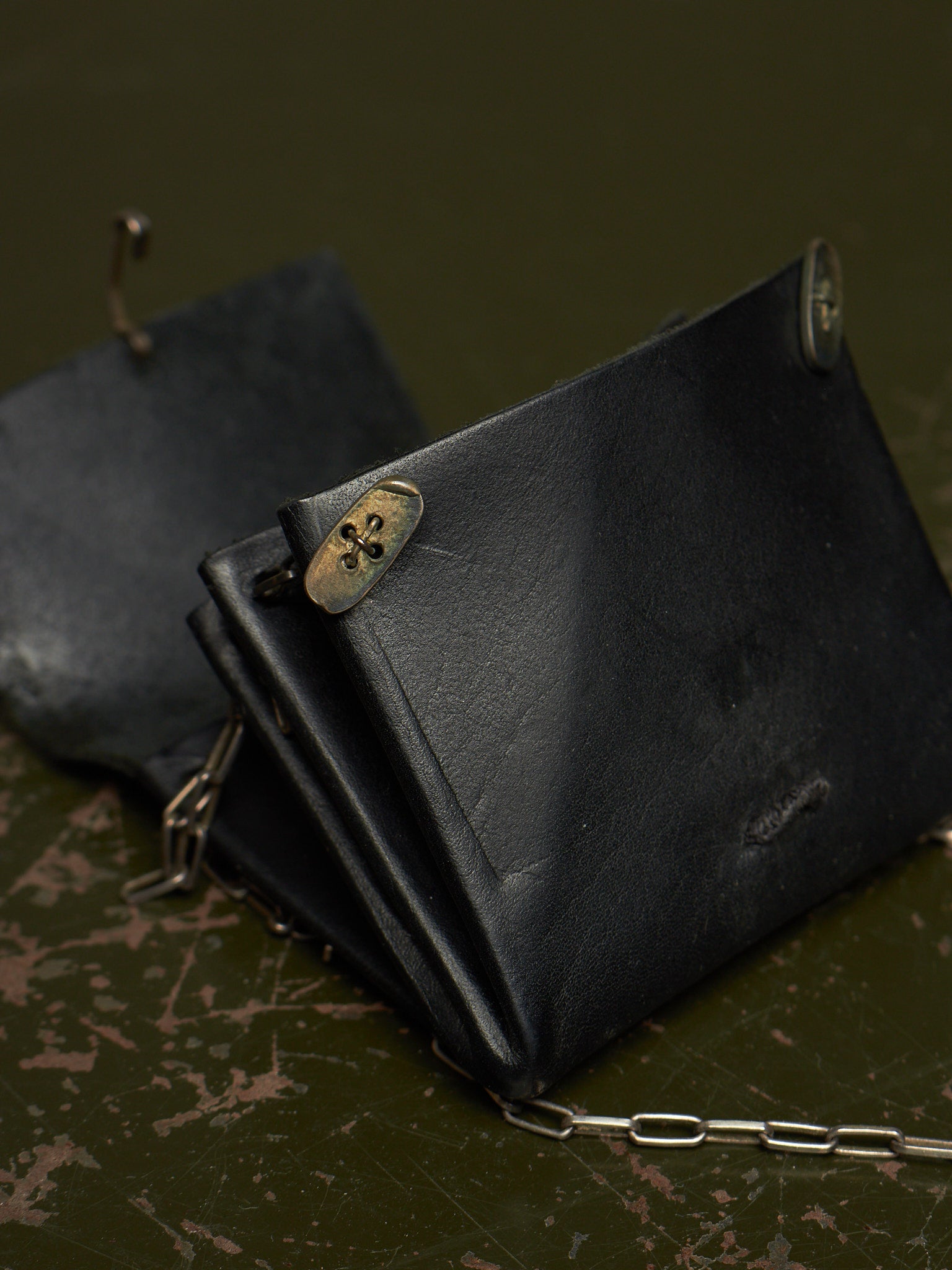 A black leather wallet with a silver chain strap and brass-tone metal clasp, photographed on a dark textured surface.