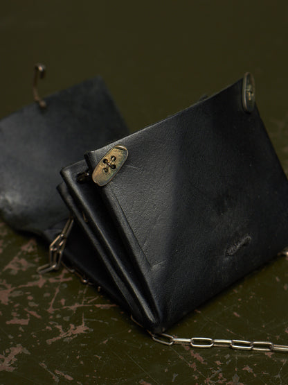 A black leather wallet with a silver chain strap and brass-tone metal clasp, photographed on a dark textured surface.